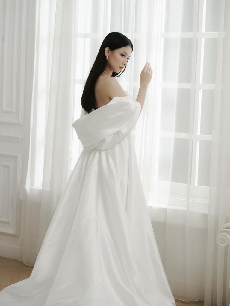Bride in an off-shoulder lace wedding gown by a bright window by CELRA